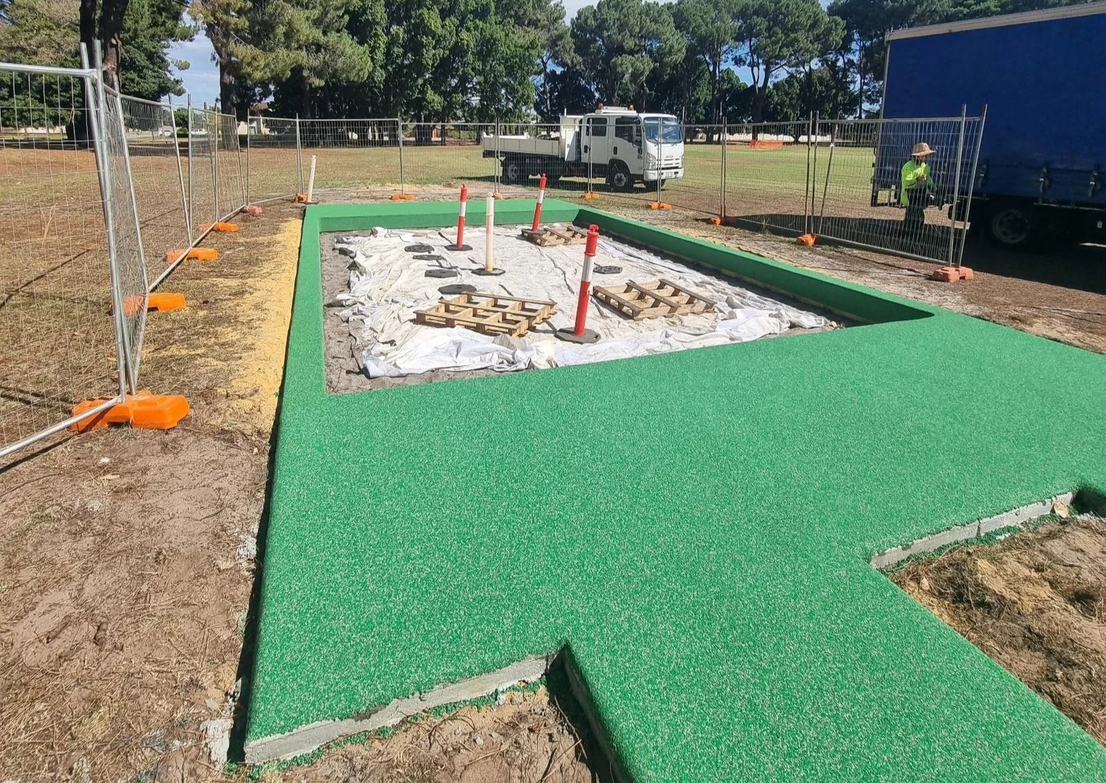 Construction site with artificial turf flooring, construction materials, orange and white safety cones, and fencing, with trucks and trees in the background.