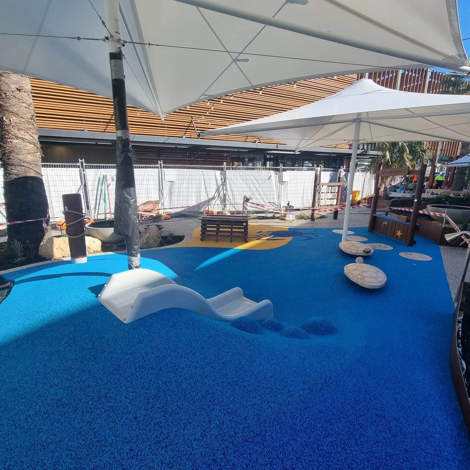 Outdoor play area with blue rubber flooring, a white slide, large umbrellas providing shade, and a wooden fence with construction work visible in the background.