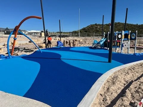 A playground with a blue rubberized surface, sand area, and play structures under construction, with a clear blue sky and hill in the background.