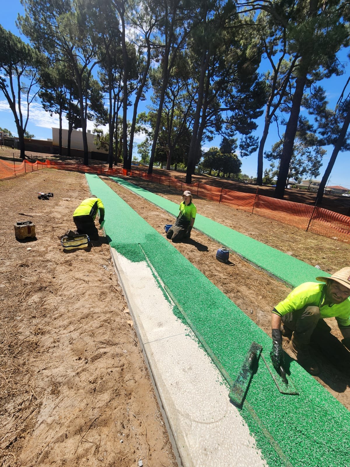 Workers painting a green pathway on a sidewalk with tools, under a clear blue sky with trees in the background.