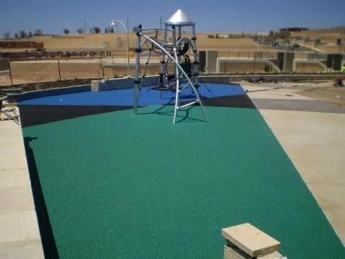 A putting green with a golf practice machine on top, set on a concrete surface outdoors, with a fence and a tent in the background.