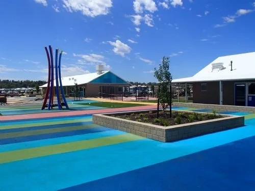 Colorful playground with a painted surface, trees in square planters, and modern buildings under a blue sky.