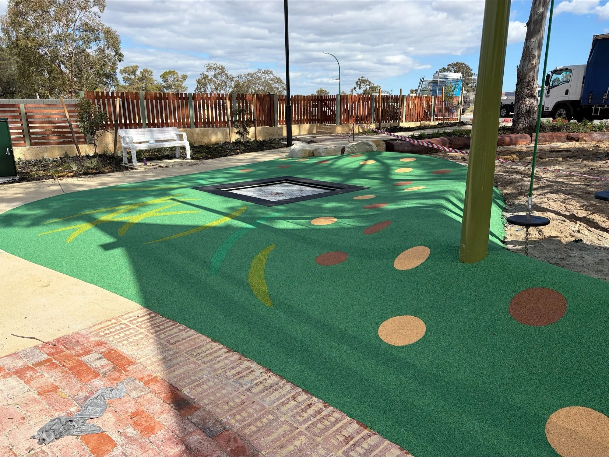 A playground area with a padded green surface decorated with colorful dots and shapes. There is a white bench, trees, a wooden fence, and construction or maintenance area with warning tape in the background. A truck and a large tree are also visible.