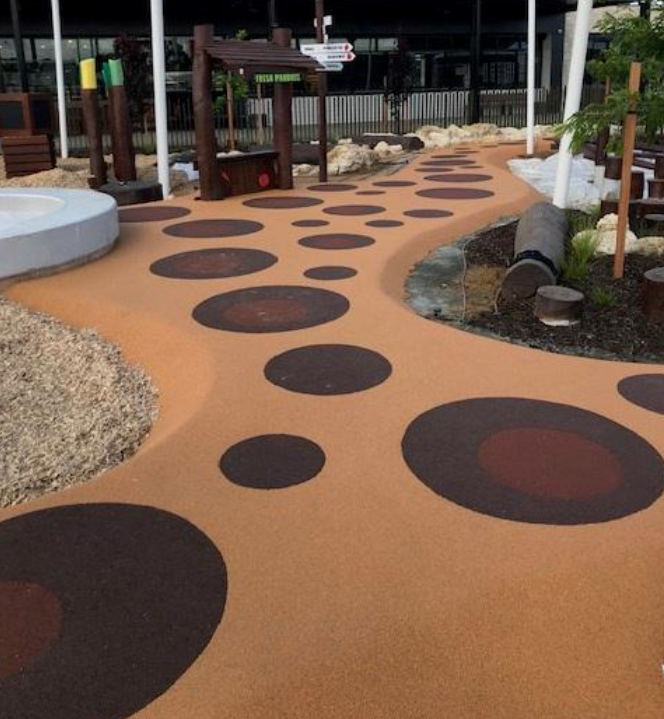 Colorful outdoor pathway with tan surface and large black and red circular patterns, surrounded by small rocks, plants, and wooden fencing.