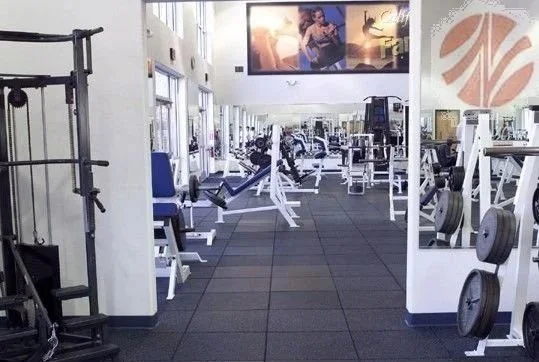 Empty gym with various workout machines and equipment, large posters on the walls, and black rubber flooring.
