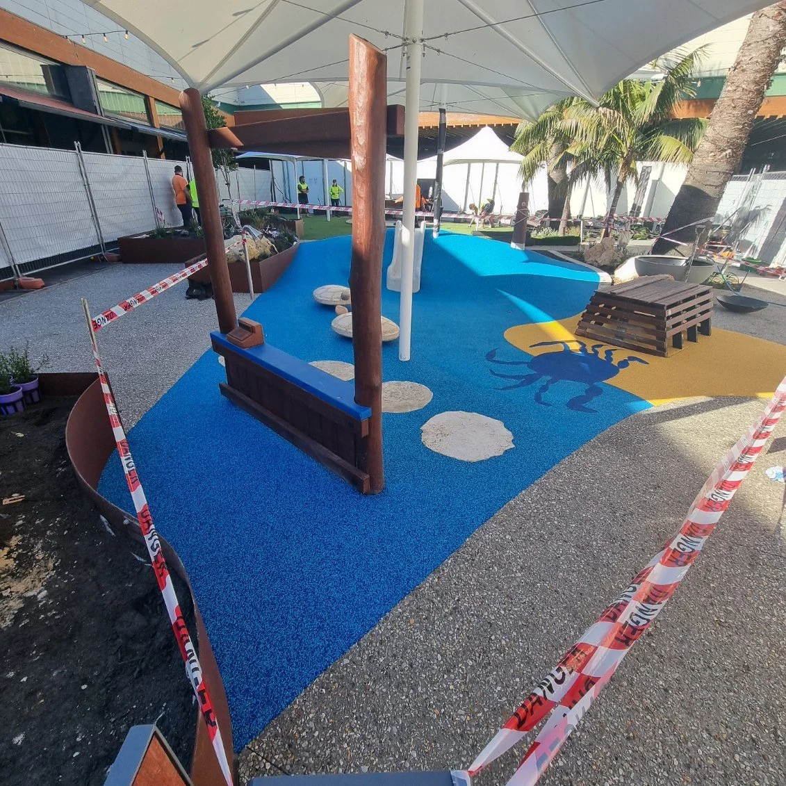 A colorful outdoor playground area under a large canopy, featuring a blue and yellow rubber surface with a lobster design, stepping stones, a wooden bench, and palm trees in the background. Construction barriers surround the space and workers are visible in the distance.