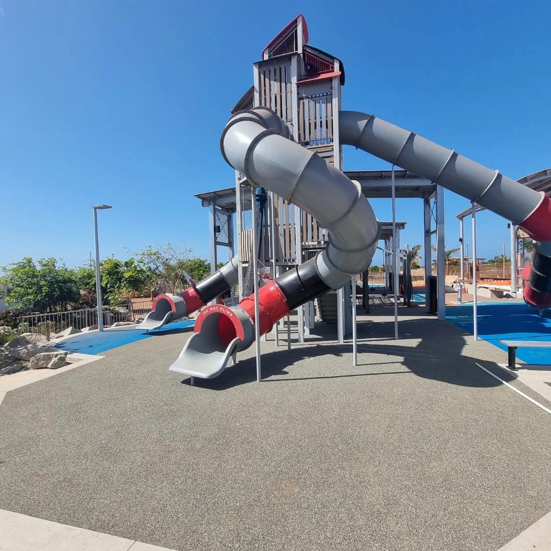 A large playground structure with multiple slides, including a long twisting tube slide. The area has a soft, rubberized surface and is outdoors with a bright blue sky.