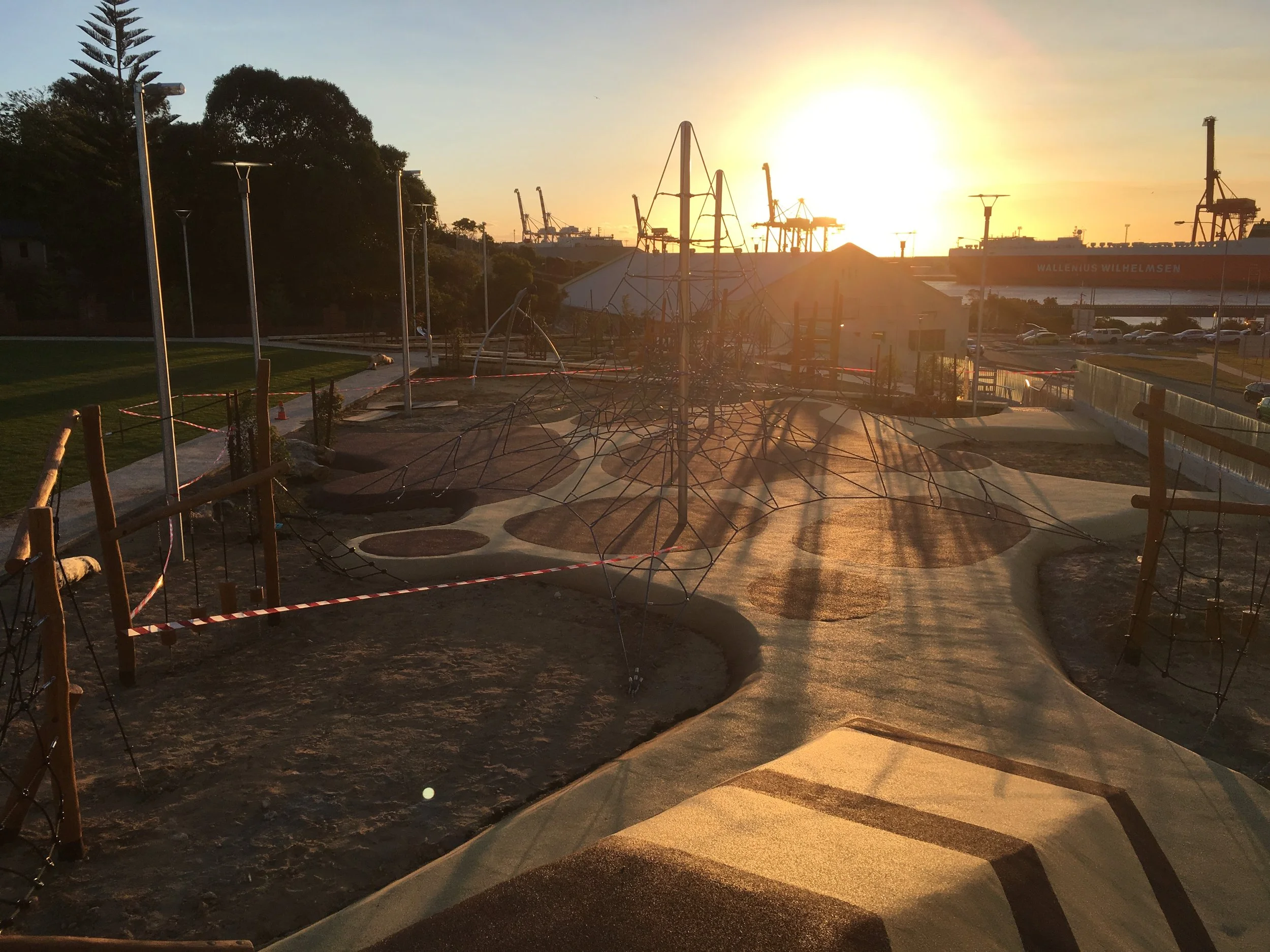 Sun setting over a waterfront park with playground equipment, trees, and a large cargo ship in the distance.