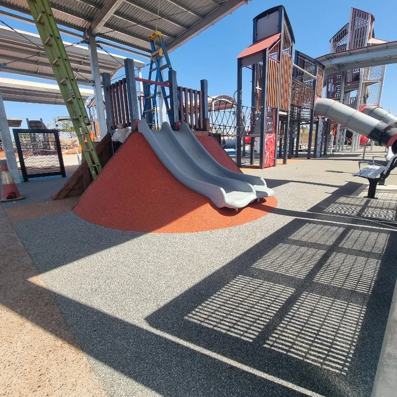 A playground with a slide, climbing structures, and slides under a metal roof, shadows cast on the ground.