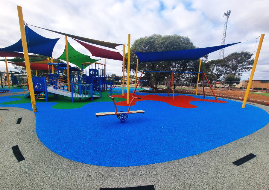 Colorful playground with shade structures, slides, swings, and a seesaw on a rubberized surface, surrounded by trees and a partly cloudy sky.