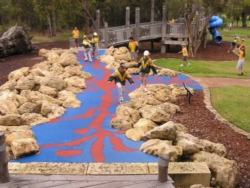 Children playing on a colorful play area with rocks, a wooden bridge, and a blue slide in a park.