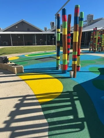 Colorful painted poles at a playground with a blue and yellow abstract rubber surface, fencing, and a house in the background.