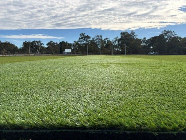 A view of a grassy sports field with a blue sky and some clouds, trees in the background, and a truck on the right side.