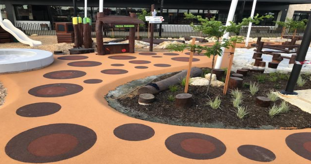 Colorful playground with a walking path featuring large painted circles, landscaped area with young trees, small plants, and wooden elements for children to play.
