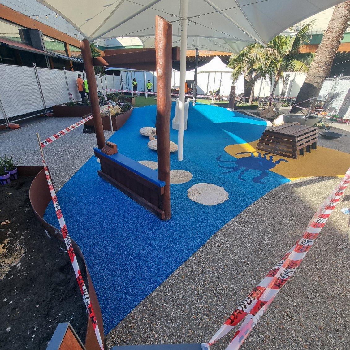 Children's outdoor playground under construction with blue and yellow rubber flooring, featuring a painted scorpion on the yellow section, and safety barriers with 'POLICE LINE DO NOT CROSS' tape around the area.