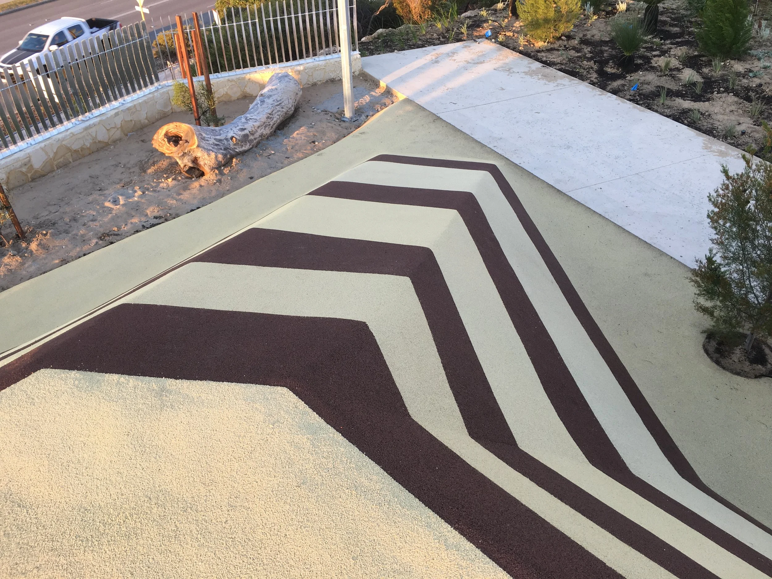 A completed landscape project, featuring a painted concrete walkway with black geometric stripes, a small garden with plants, and a section under construction with a tree trunk and dirt. There is a black metal fence and a nearby road with a parked ve