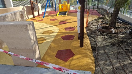 Children's playground with colorful rubber flooring, a swing set, and various play structures, surrounded by a fence and some trees.
