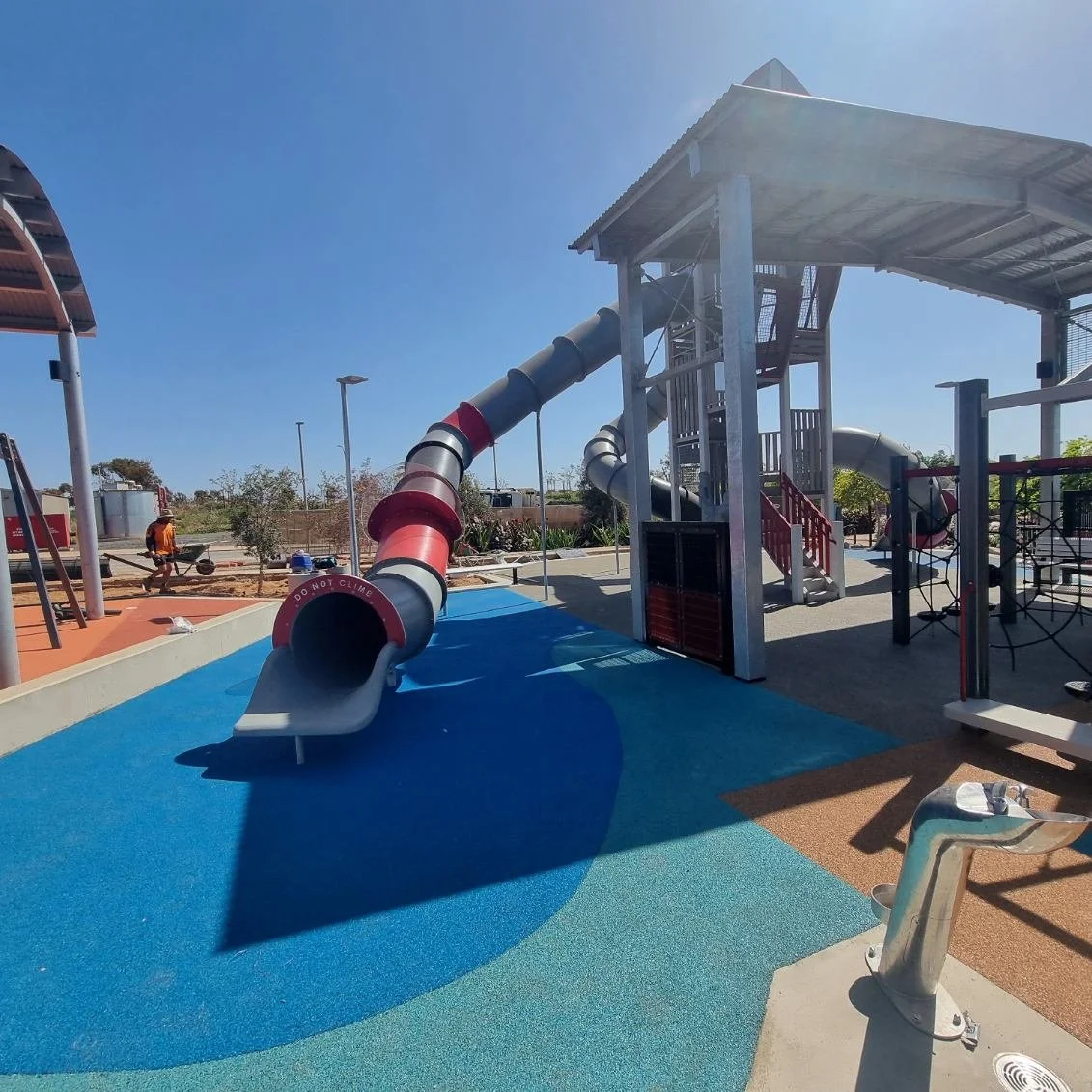 Empty playground with a large red and gray tube slide, covered climbing structure, and a water fountain in the foreground, under clear blue skies.