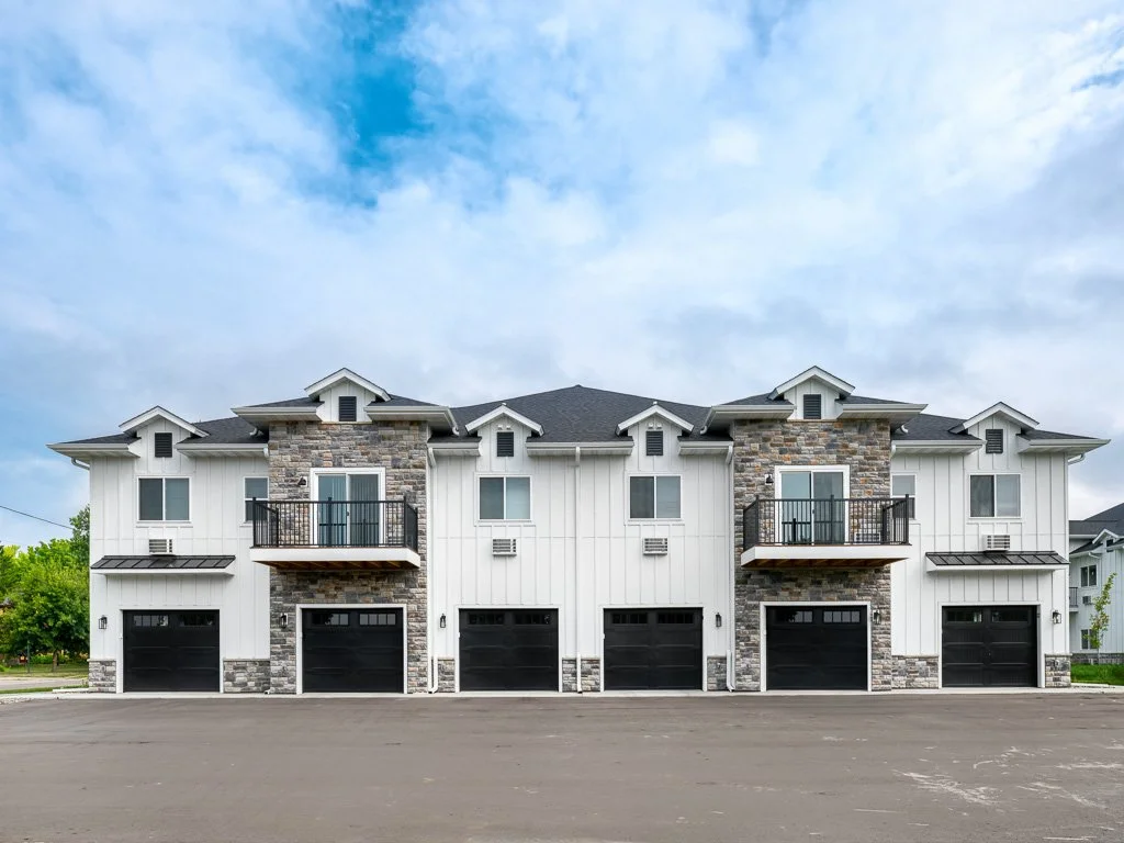 A modern multi-unit residential building with black garage doors, white exterior, stone accents, and small balconies, under a partly cloudy sky.