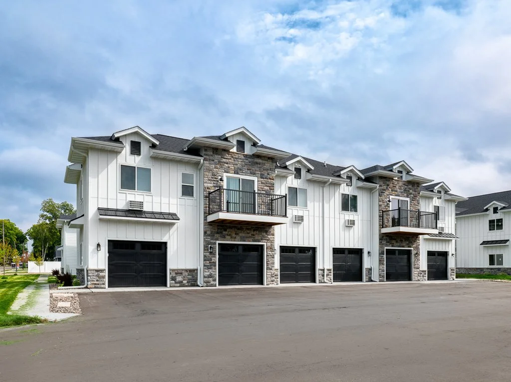 A row of modern townhouses with white siding, stone accents, black garage doors, and small balconies under a partly cloudy sky.