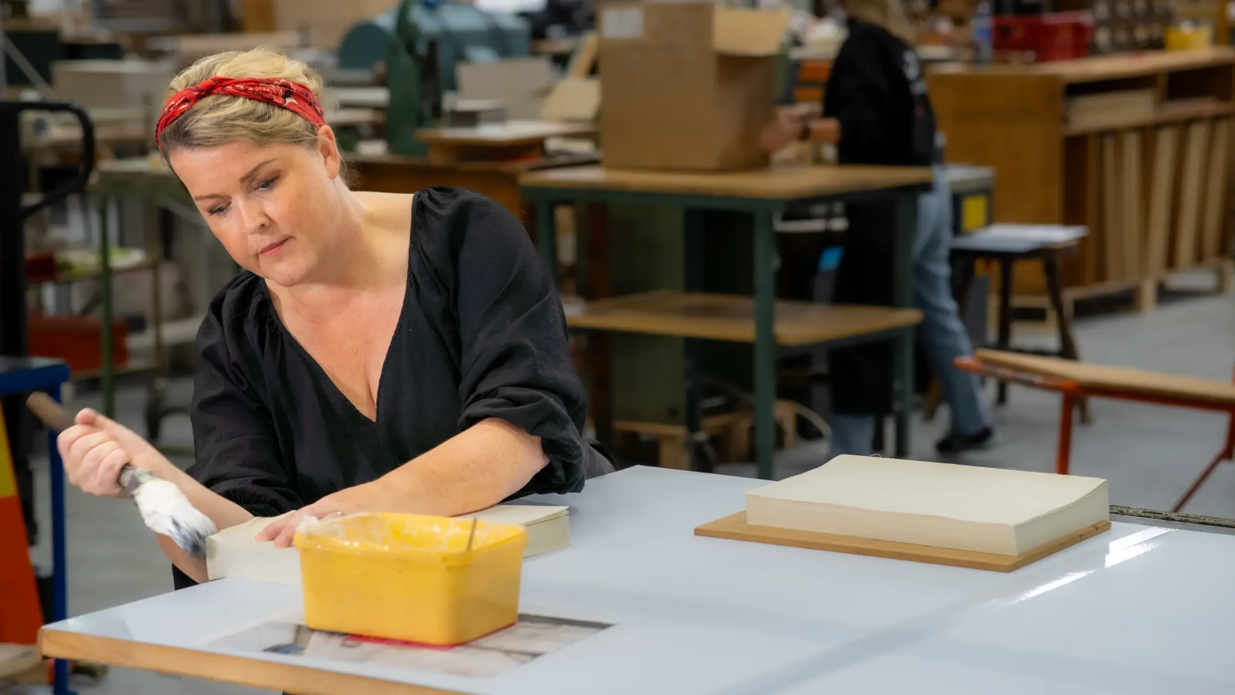 En kvinne med blondt hår og rød skjerf jobber i et verksted eller på en fabrikk, omgitt av arbeidsbord, verktøy og materialer.
