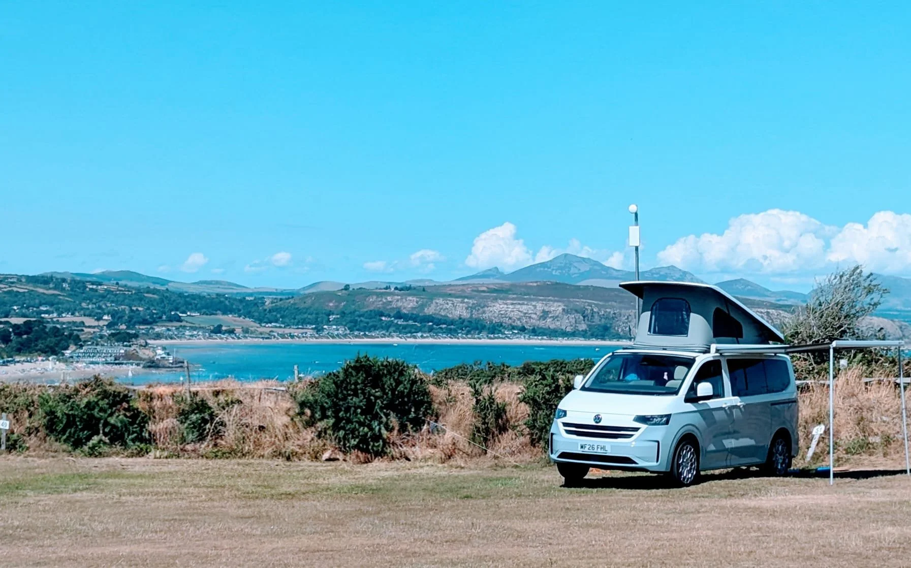 A white camper van with a pop-up roof parked on a grassy area near water, with hills and mountains in the background, on a sunny day with a blue sky.