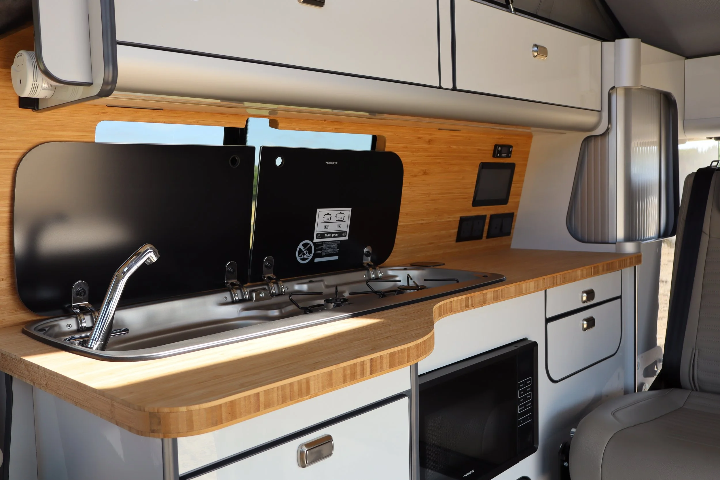 Interior of a compact camper van kitchen with a wooden countertop, sink, stove, and cabinets, along with a small TV or monitor and electrical outlets.
