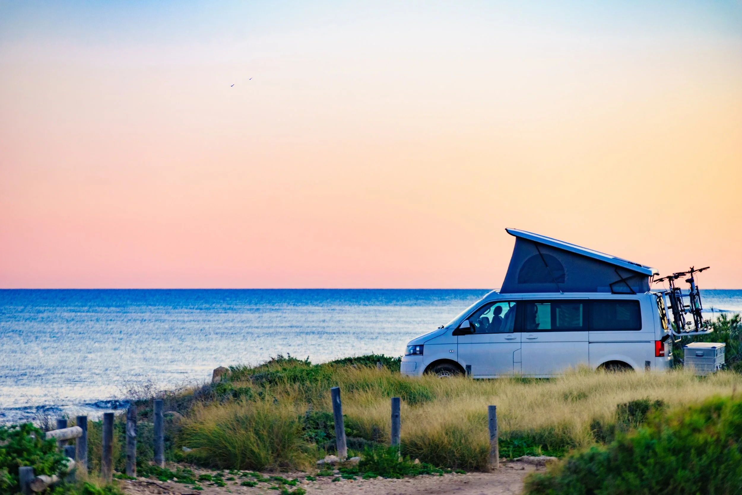 A white camper van parked on a grassy area near the ocean with a bike rack on the back and a pop-up roof, beneath a pastel-colored sky at sunset.