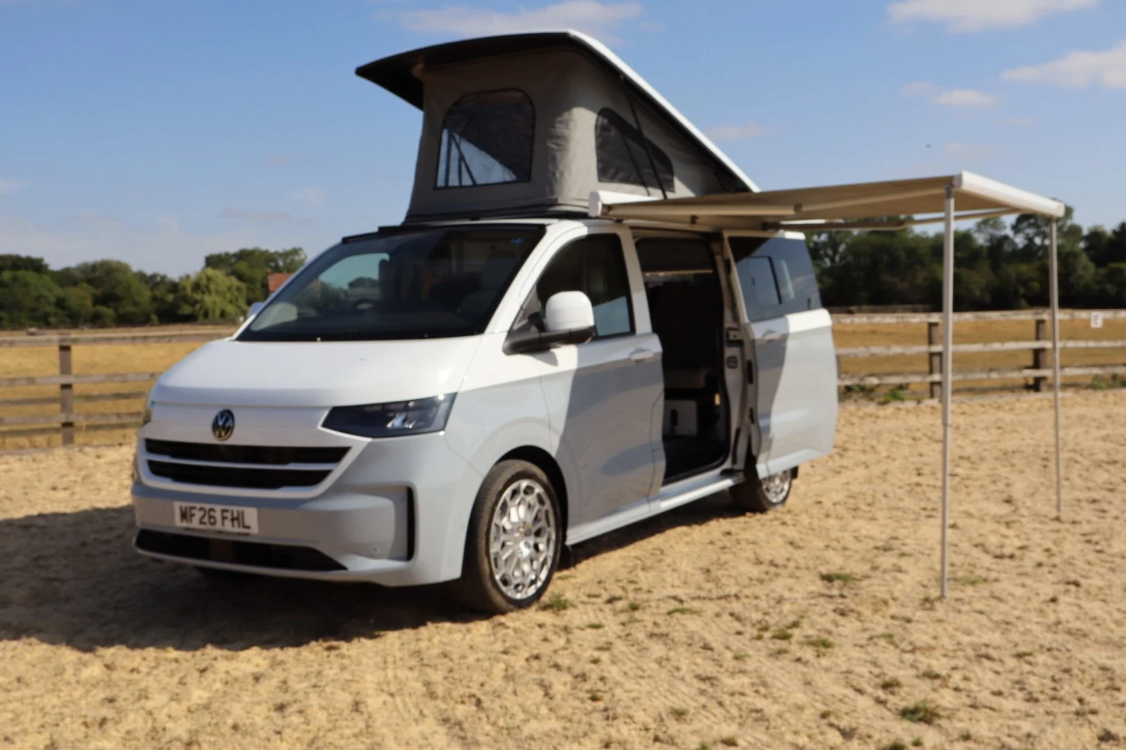 A white camper van with a pop-up roof parked on a sandy area with a wooden fence and trees in the background.
