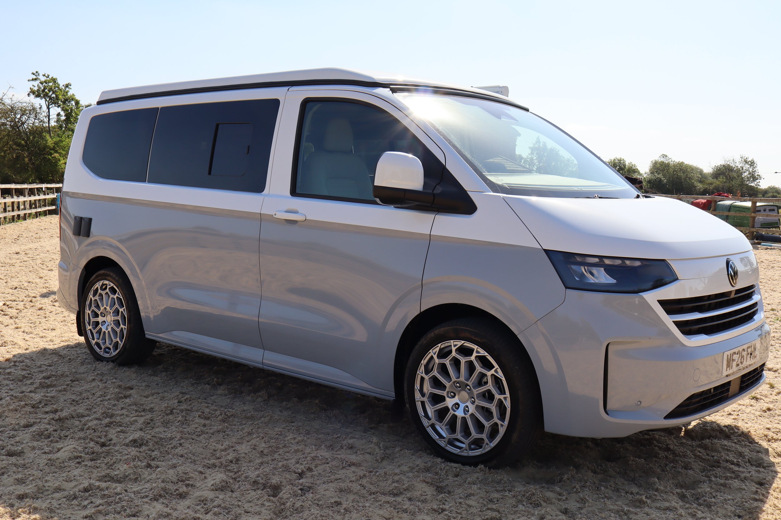 White modern van parked on sandy ground with trees and a fence in the background under clear sky.