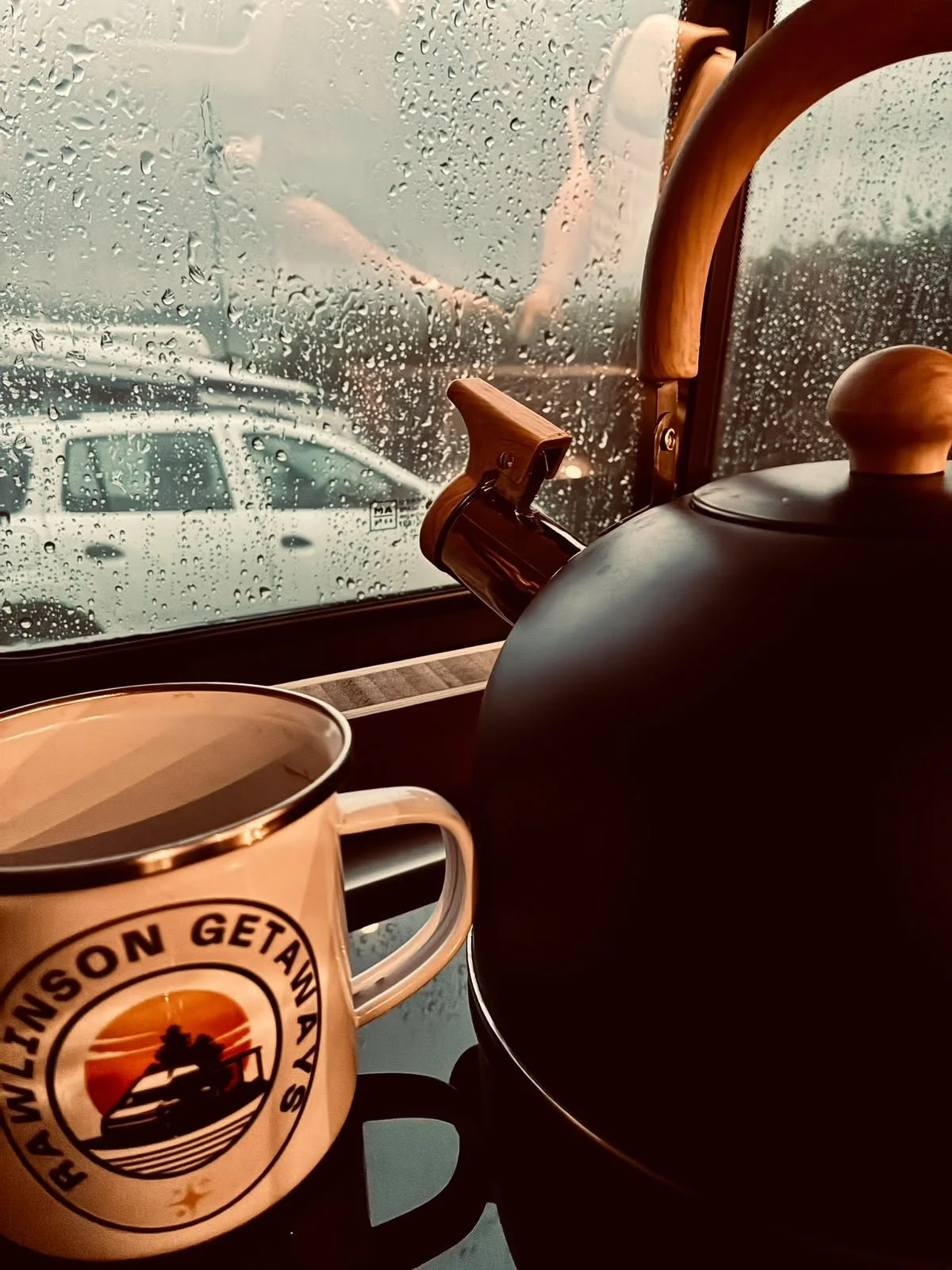 A rainy scene outside a window with water droplets on the glass. Inside, there is a stovetop kettle, a mug with a logo, and a coffee pot. The mug has a mountain and sunset design with the text 'Wisconsin Getaway'.