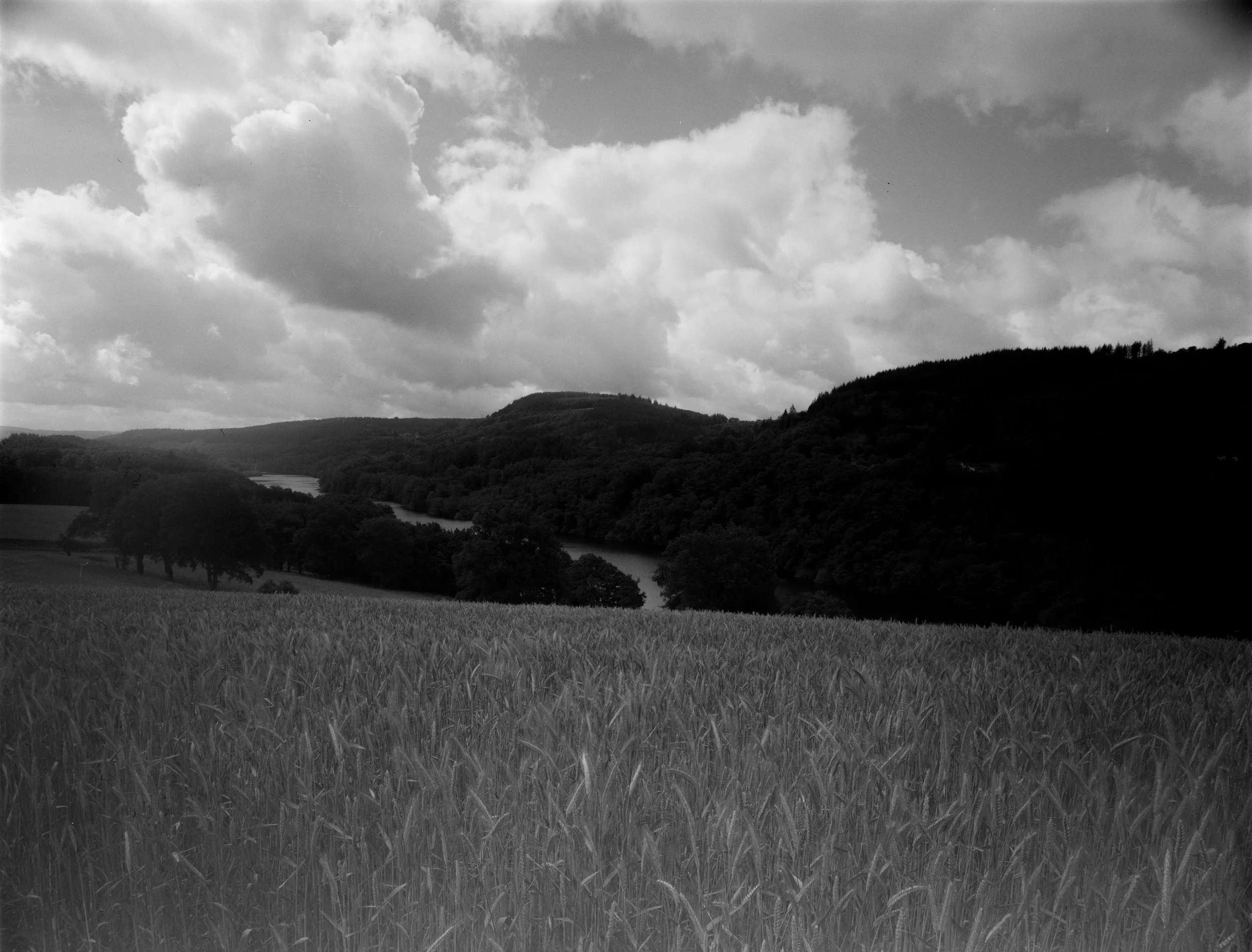 A black and white landscape photo featuring rolling hills, a river, and a dramatic cloudy sky.
