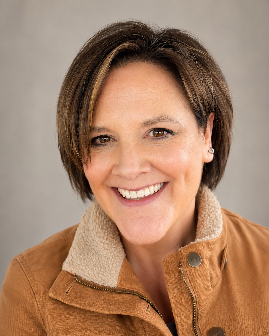 A woman with short brown hair smiling at the camera, wearing a brown jacket with a shearling collar.