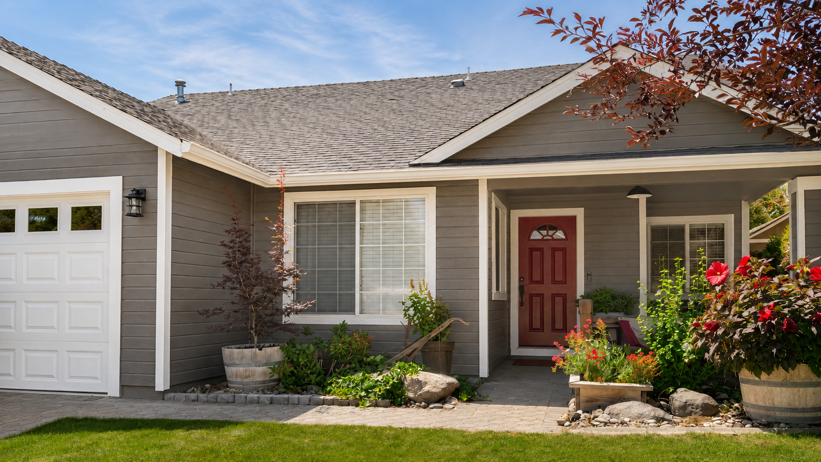 Front view of a suburban house with a gray exterior, red front door, white trim, and a landscaped garden with potted plants and blooming flowers.