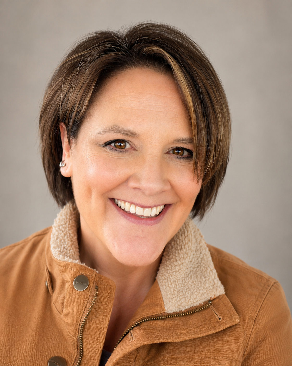 A woman with short brown hair, brown eyes, and fair skin smiling at the camera, wearing a tan jacket with a fleece collar and small earrings, against a neutral background.
