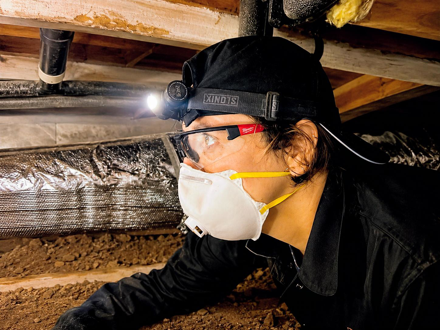 A person working underground with safety gear, including a headlamp, face mask, glasses, and black clothing, during an excavation or construction project.