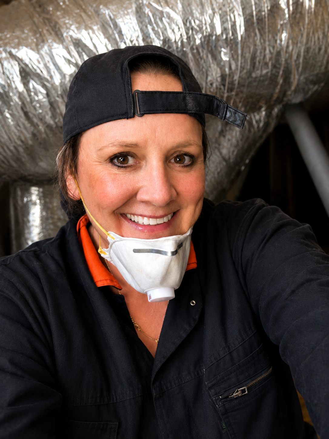A woman taking a selfie inside an industrial or ventilation setting, wearing a black cap backwards, a face mask pushed down on her chin, and a dark work jacket.