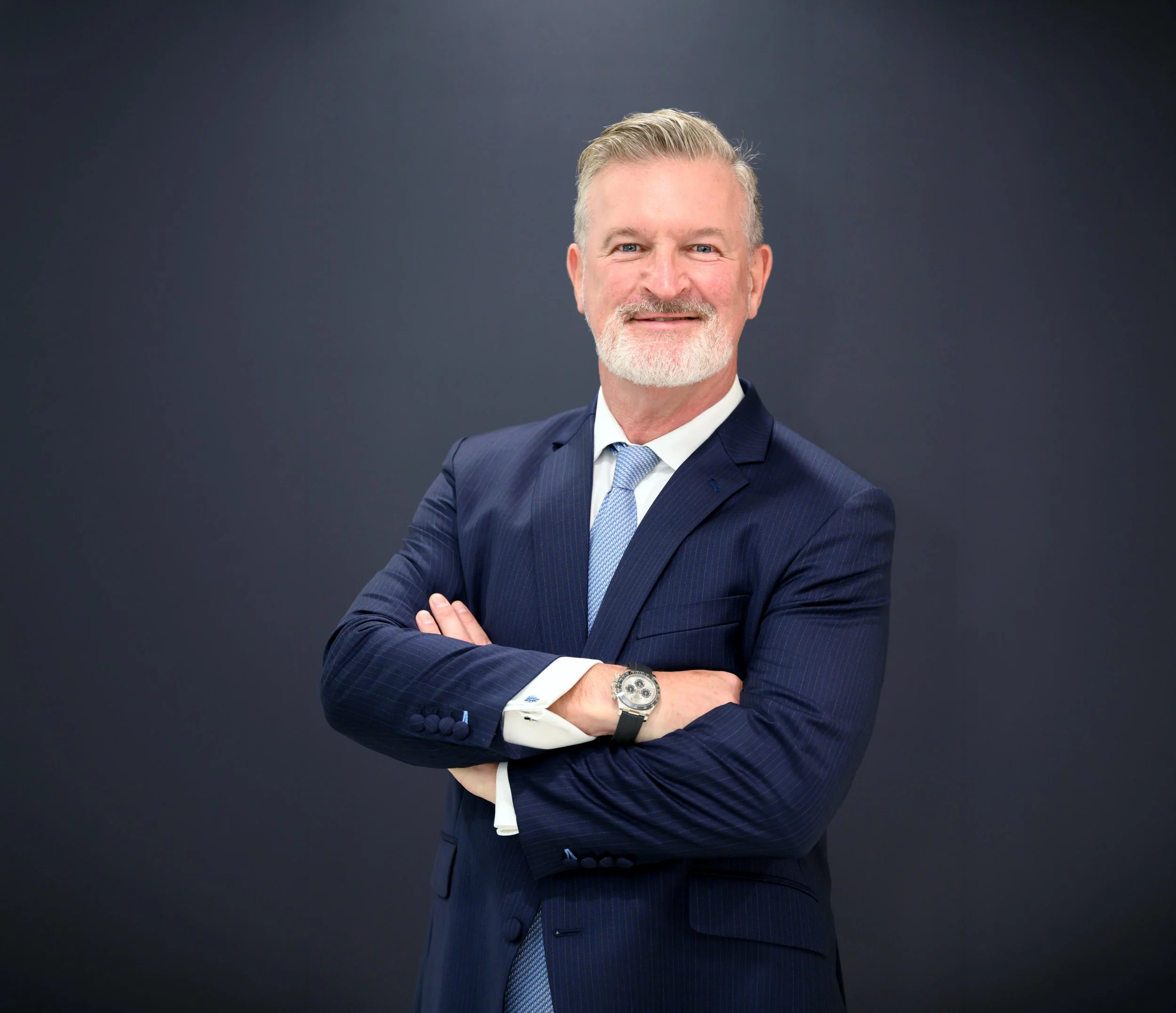 A middle-aged white man stands at three quarter view. He has his arms gently crossed. He wears a navy pin-striped suit, white short, and light blue striped tie. He has grey hair and a short white beard. He stands against a dark studio background.
