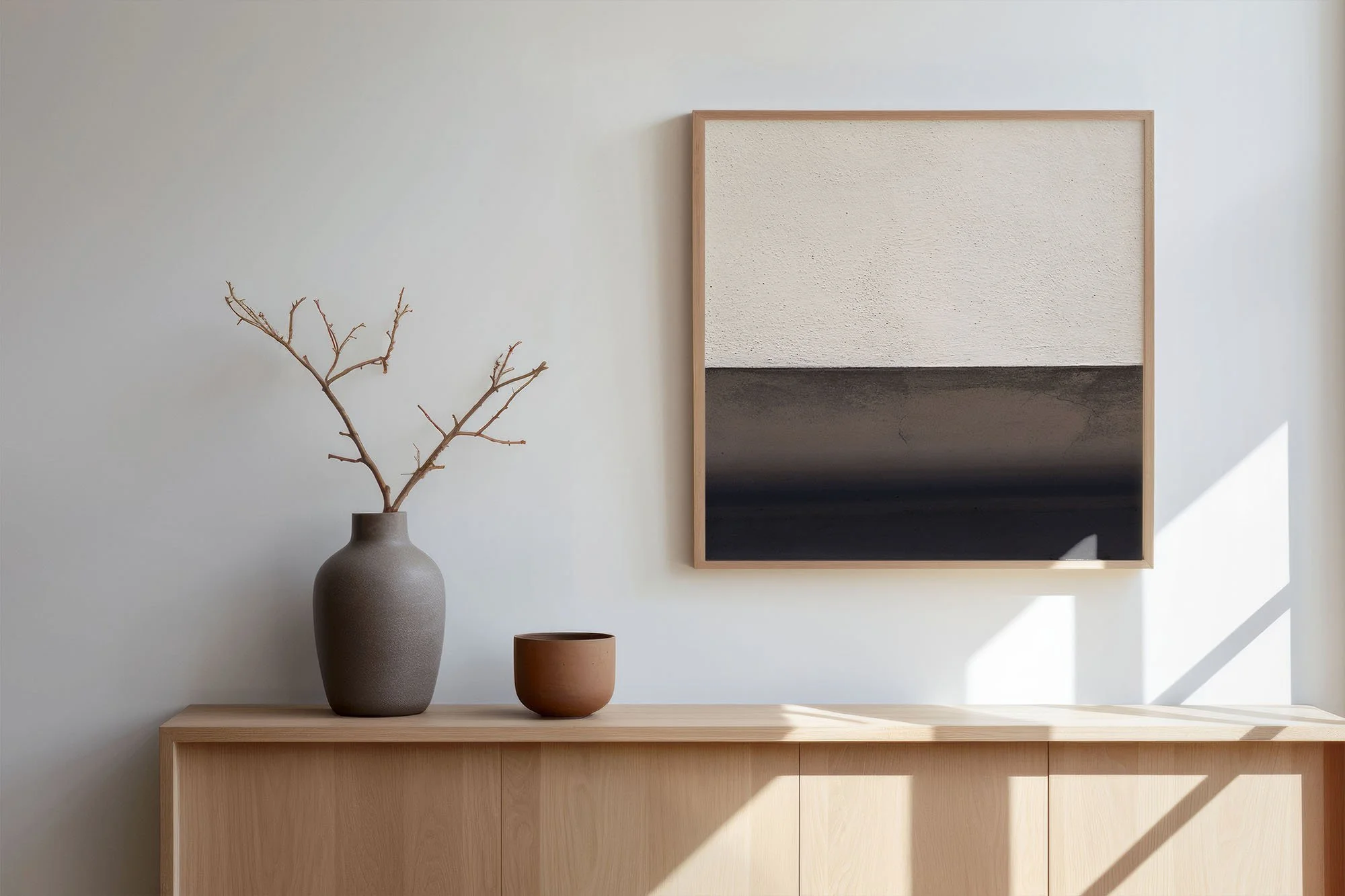 Vaso con ramo secco, due ciotole in ceramica e quadro astratto sui toni del marrone su parete bianca, con mobili in legno chiaro.