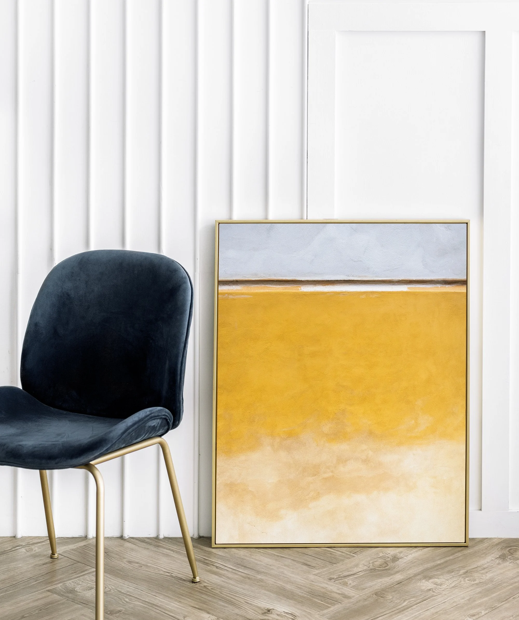 Sedia blu in velluto con gambe d'oro e quadro con sfumature di giallo dietro. Sfondo con pannelli di legno e parete bianca.
