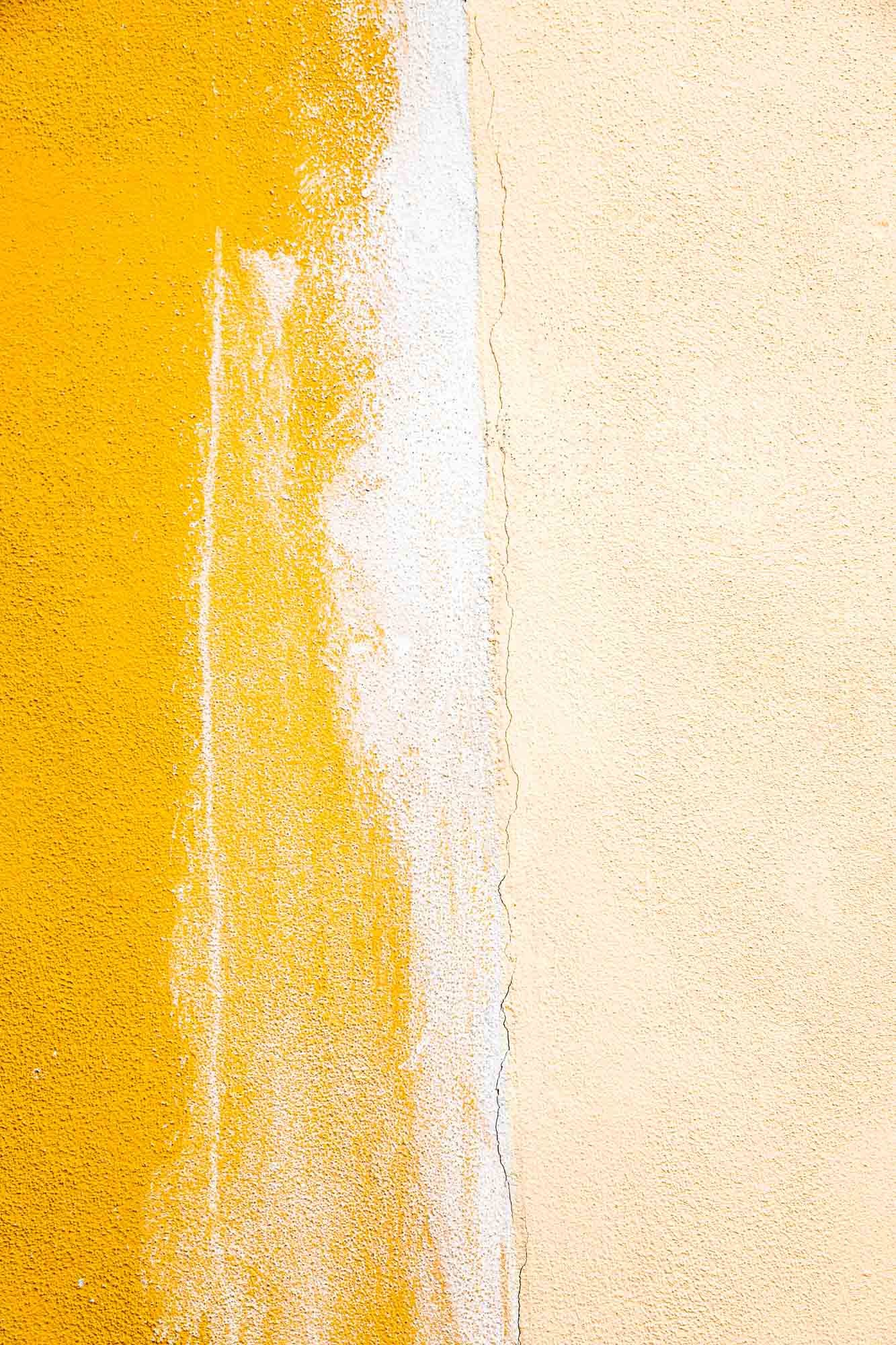 Parete con vernice gialla che si sfalda sulla sinistra e vernice beige liscia sulla destra, con una linea di separazione irregolare e una crepa centrale.