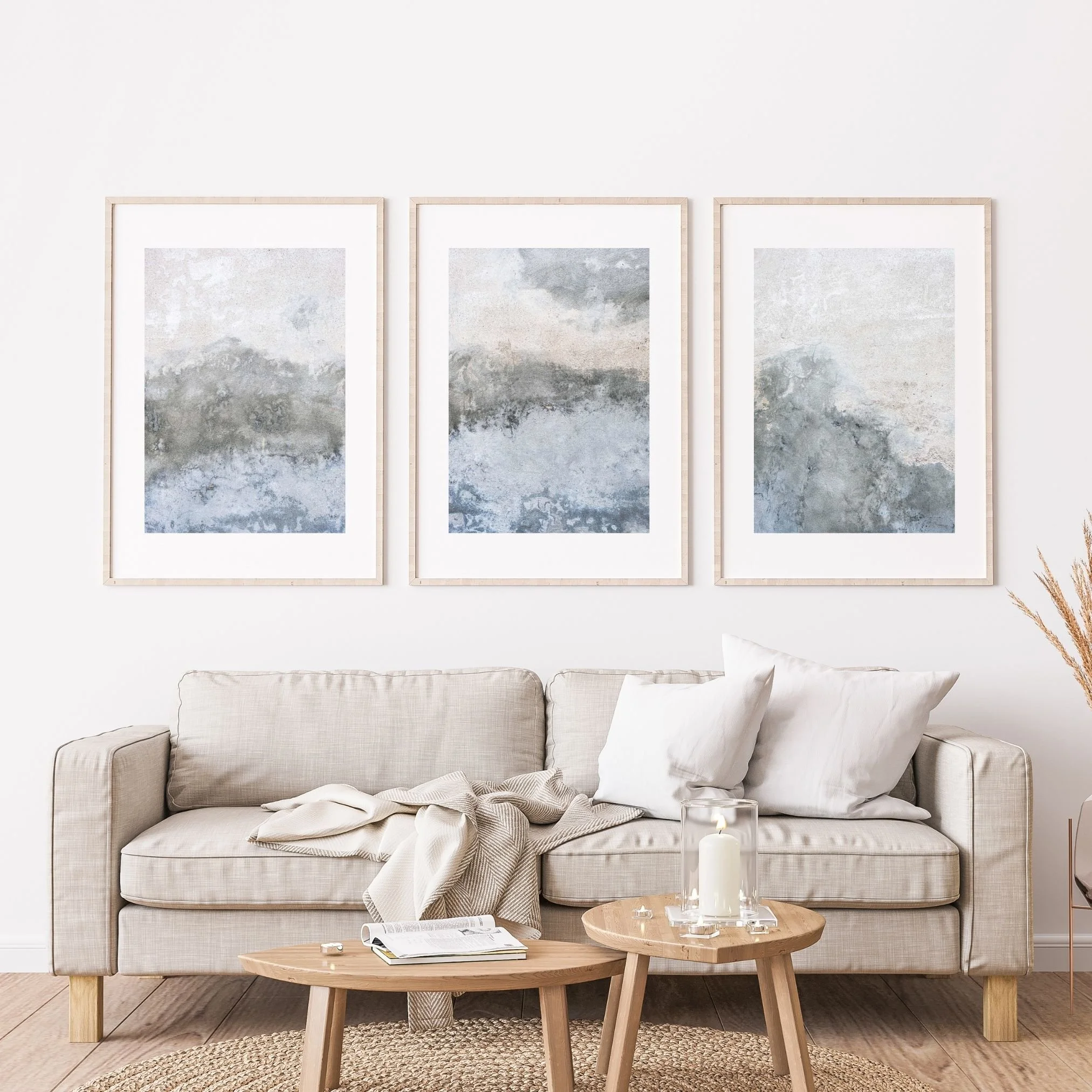 Soggiorno con divano beige, cuscini, tavolini in legno, libri, candle, decorazioni naturali, tre quadri astratti su parete bianca.