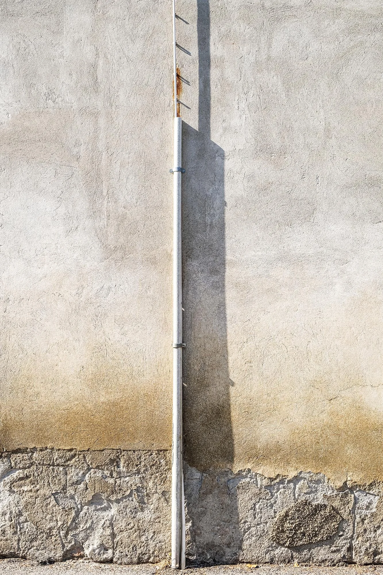 Muro di cemento, con un tubo bianco verticale e una staffa metallica che lo sostiene, e un'ombra del tubo sul muro.