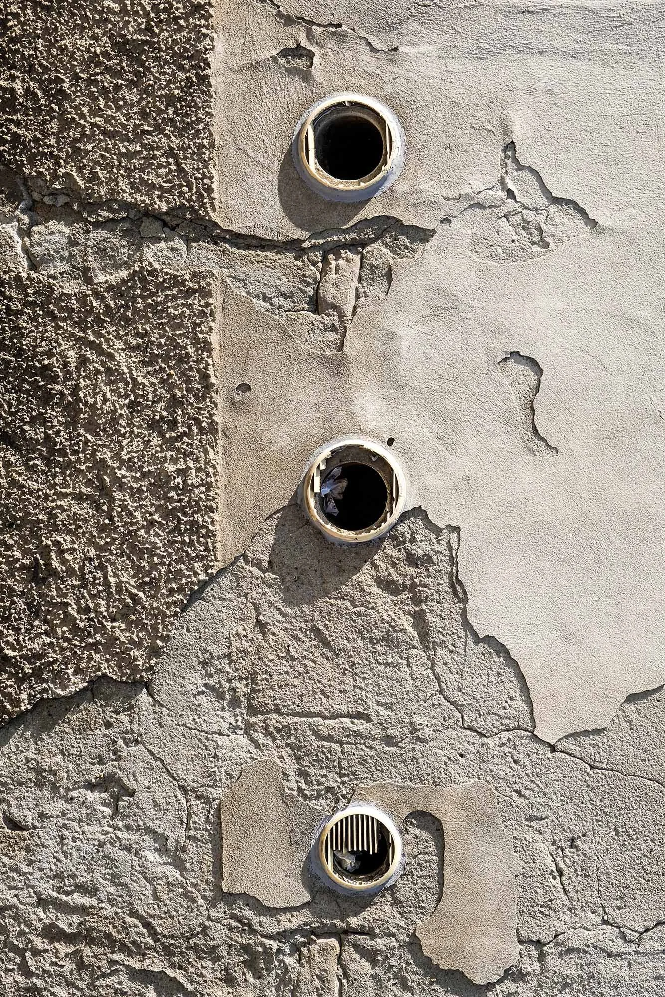 Parete in cemento con superficie rotta e tre prese di aria o ventilazione, di colore beige, con alcuni oggetti all'interno.