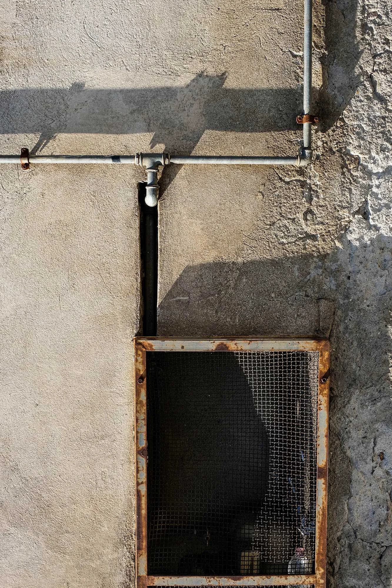 Un muro con tubi metallici e una griglia arrugginita, sotto un'apertura rettangolare.