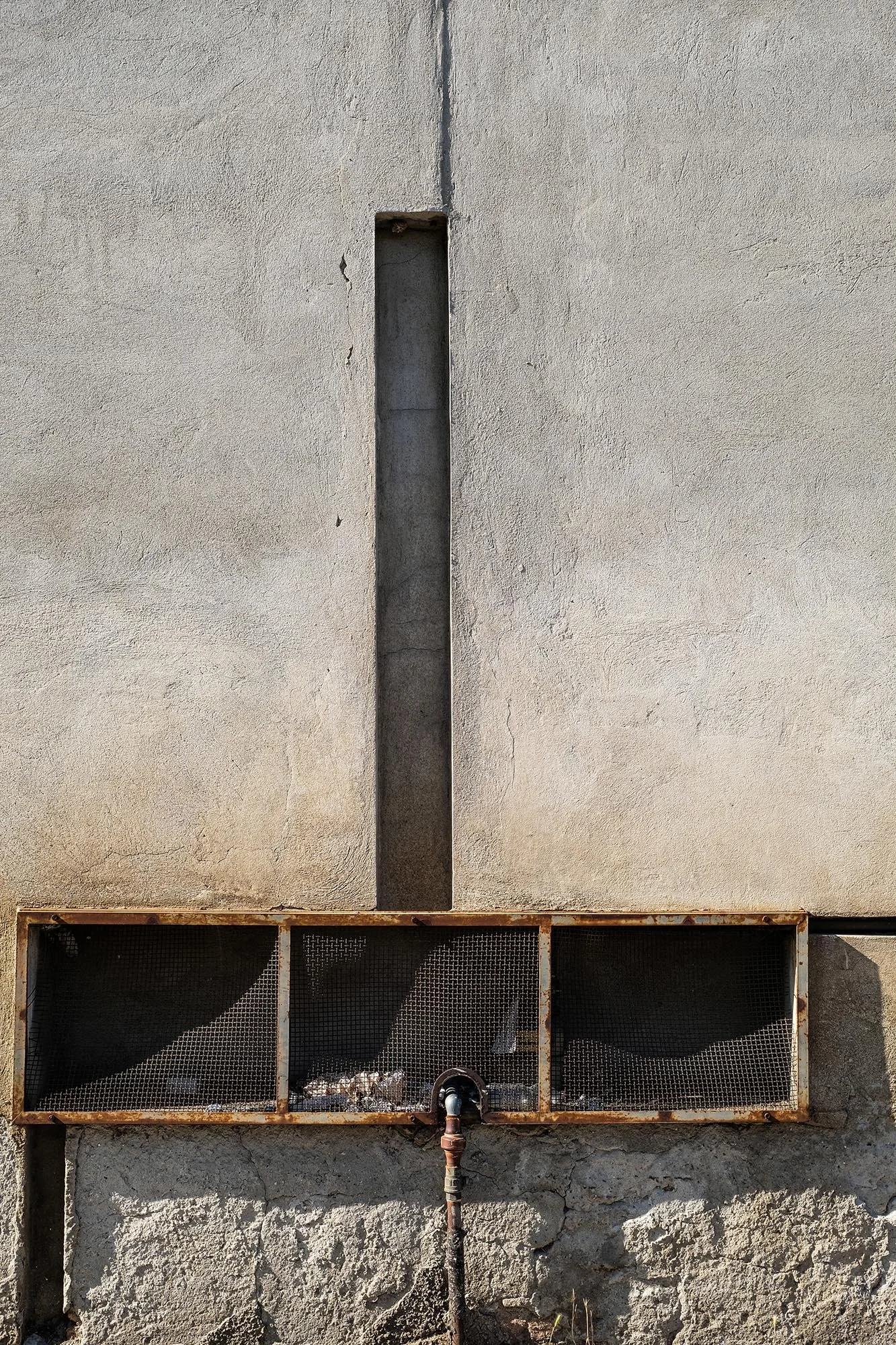 Un muro di cemento con una fessura verticale e una struttura metallica sottostante. La struttura appare arrugginita e contiene alcuni rifiuti di plastica. La superficie del muro è segnato da crepe e danni.