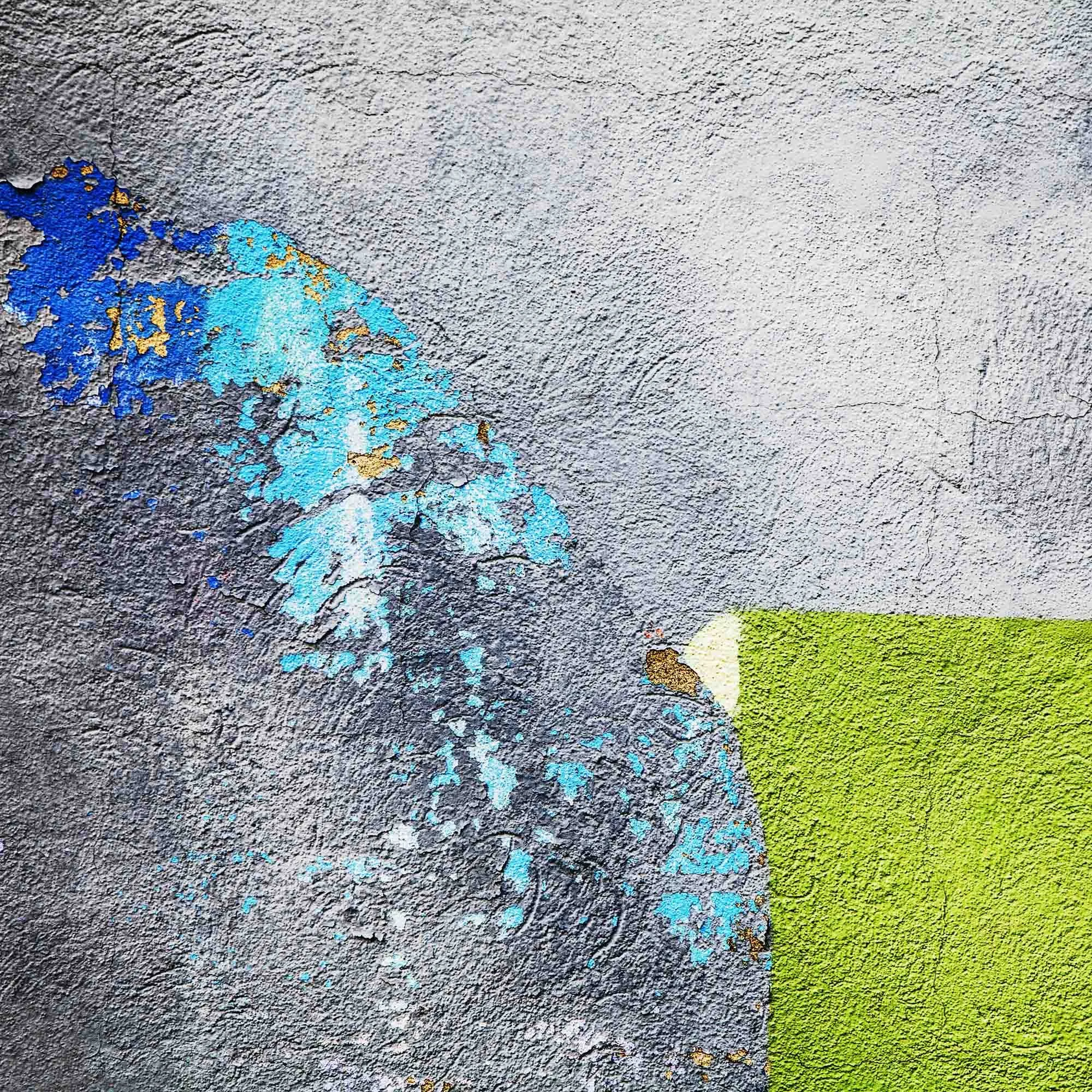 Superficie di un muro grigio con vernice in parte sfaldata che rivela colori vivaci come blu, verde e giallo sotto la superficie.