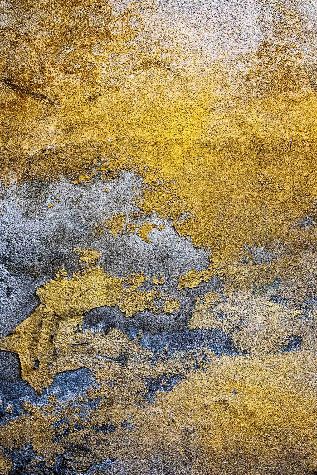 Superficie di muro con vernice gialla che si sfalda e mostra uno strato sottostante di vernice grigia.