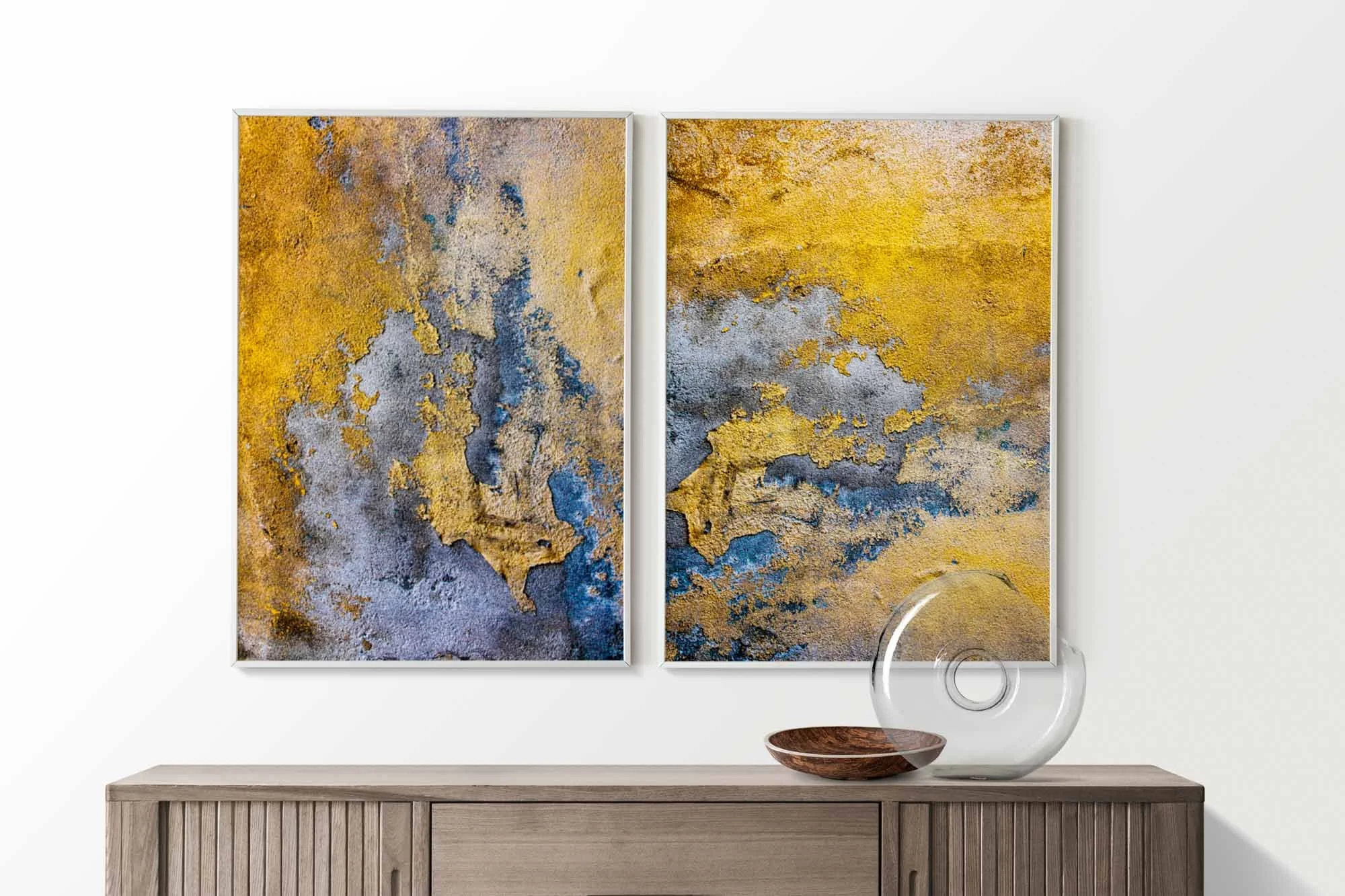 Due quadri astratti con superfici ruvide in tonalità di giallo, blu e grigio. Sono appesi su una parete bianca sopra un mobile in legno con un vaso di vetro e un piatto di legno.