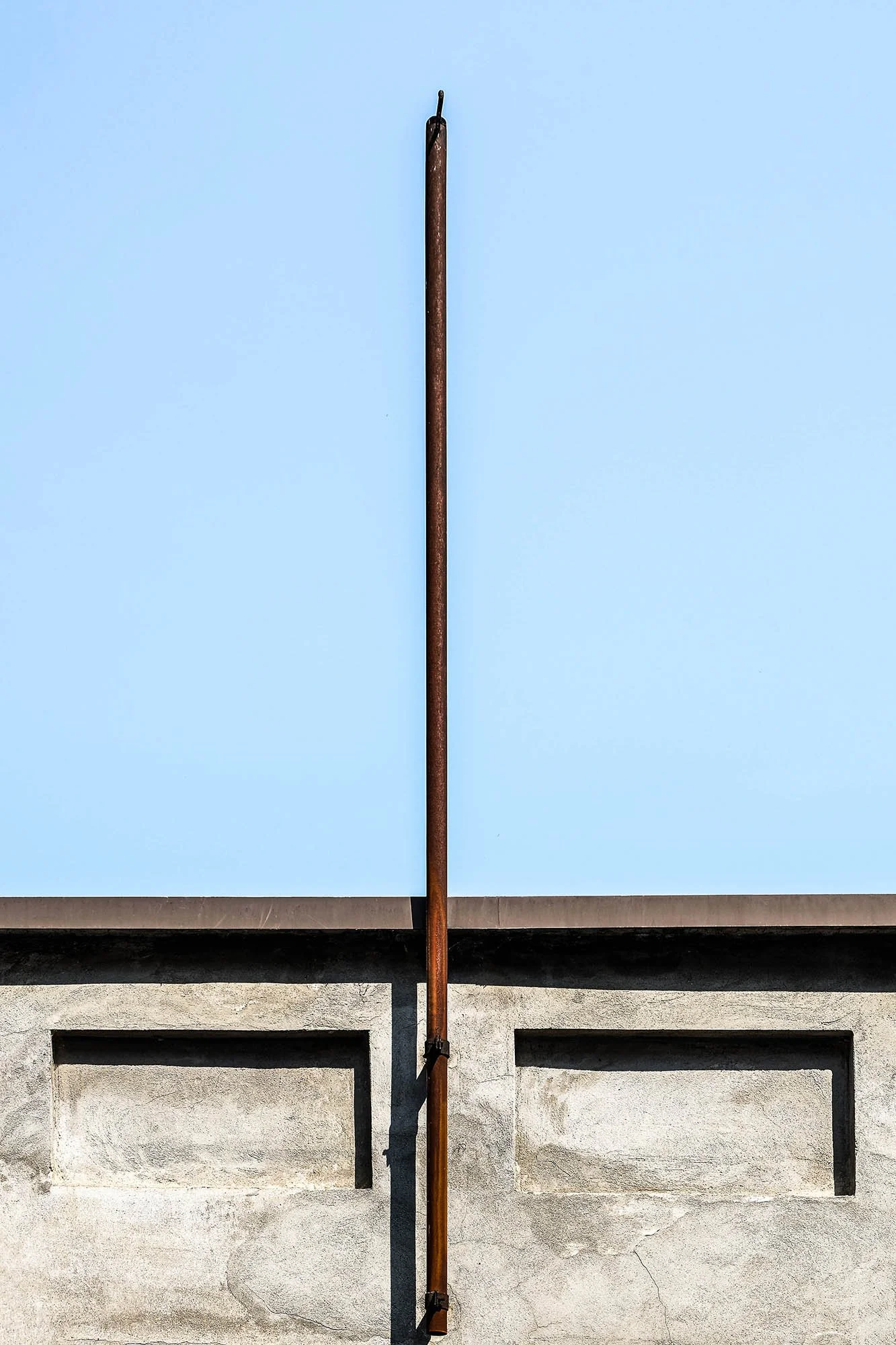 Una parete grigia con una striscia di cemento in basso, sulla quale è fissata una tubazione di ferro arrugginita che si estende verticalmente verso il cielo.